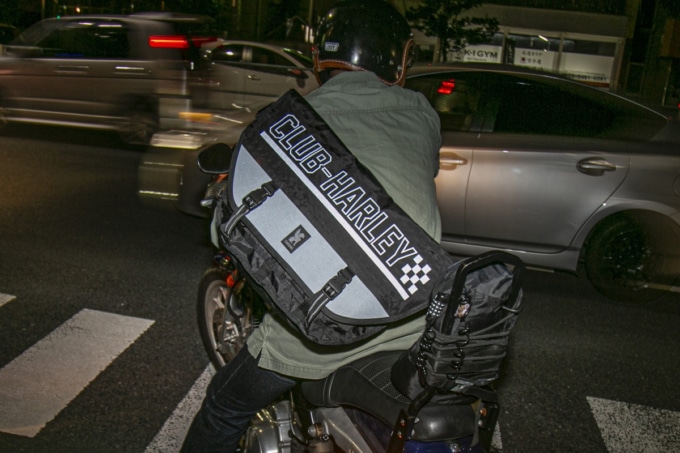 走行中にバッグを背負うシーン:夜の道路を走行中のライダーが背負うバッグのリフレクターがヘッドライトの光を反射し、視認性を高めている様子。荷物が揺れないようボディにフィットしている。