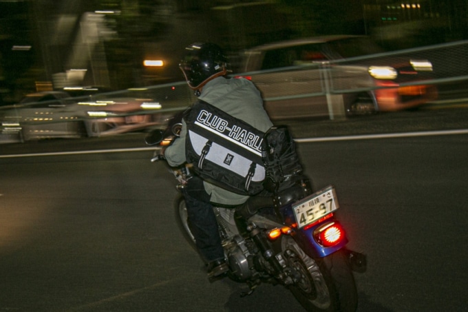 バイクに跨りバッグを背負うライダー:背中に斜め掛けされたコラボメッセンジャーバッグを背負ってハーレーに跨るライダー。大きく描かれた「CLUB‑HARLEY」のロゴとチェック柄が際立ち、バイクとバッグの一体感が印象的。
